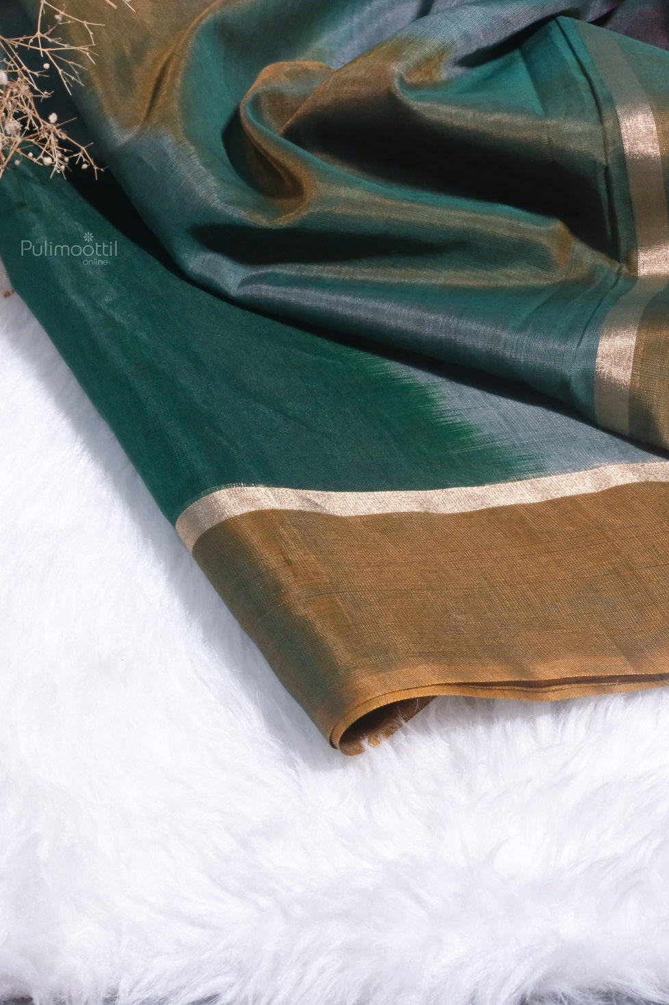 A detailed shot of the saree’s end piece, showcasing deep green hues blended with golden mustard and highlighted by a subtle gold zari border. The fabric lies on a white furry surface.