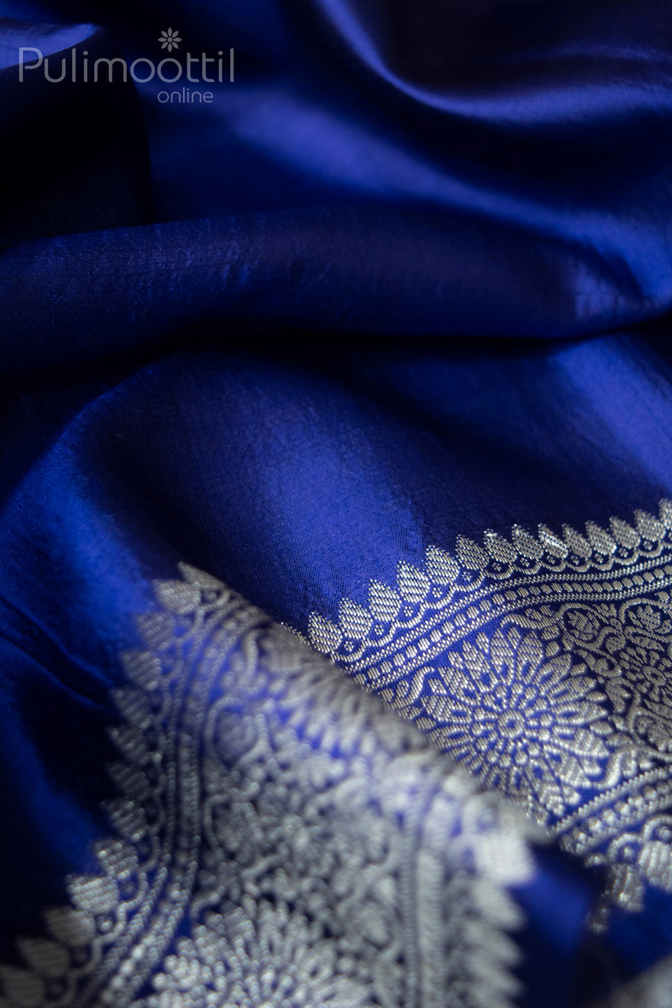 Soft focus close-up of navy blue Banarasi silk saree featuring intricate gold and silver zari pallu with floral brocade design on lustrous silk fabric.

