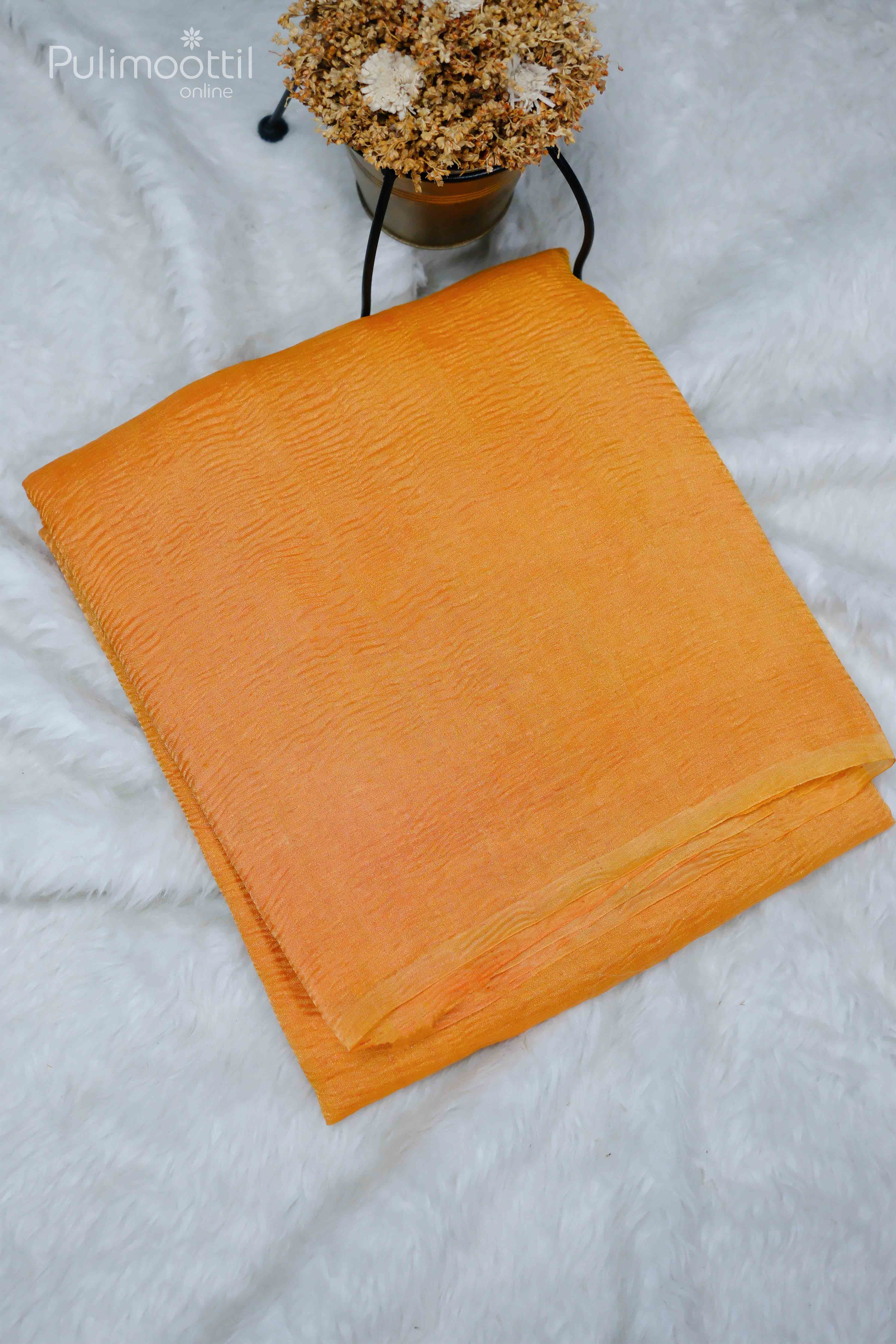 Neon orange crushed tissue saree neatly folded, plain without border, placed on a soft white surface with a potted plant in the background.