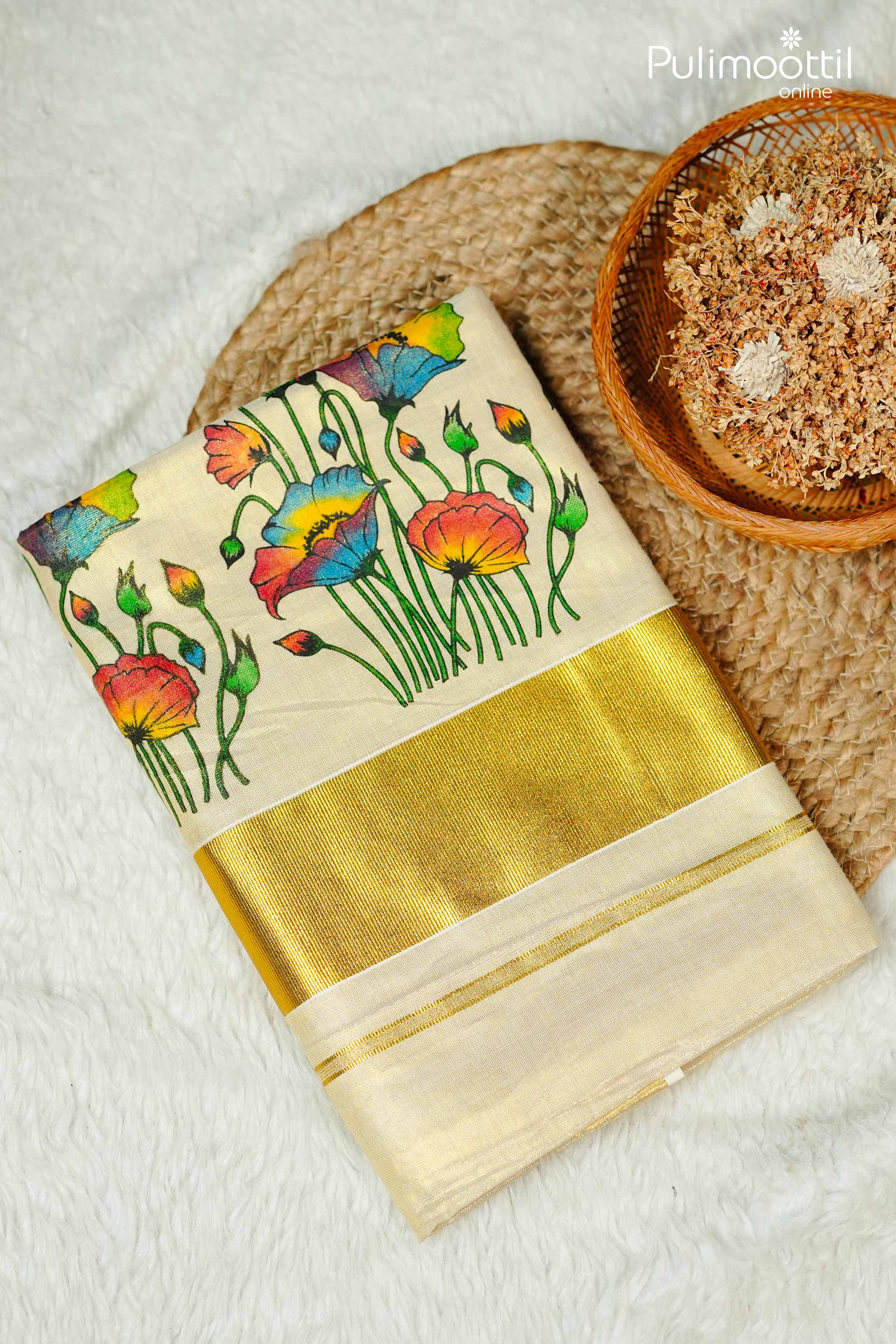 Off-white Kerala set saree with vibrant multicolor floral print on body and golden tissue border, neatly folded on a textured mat with dried flowers in a basket.