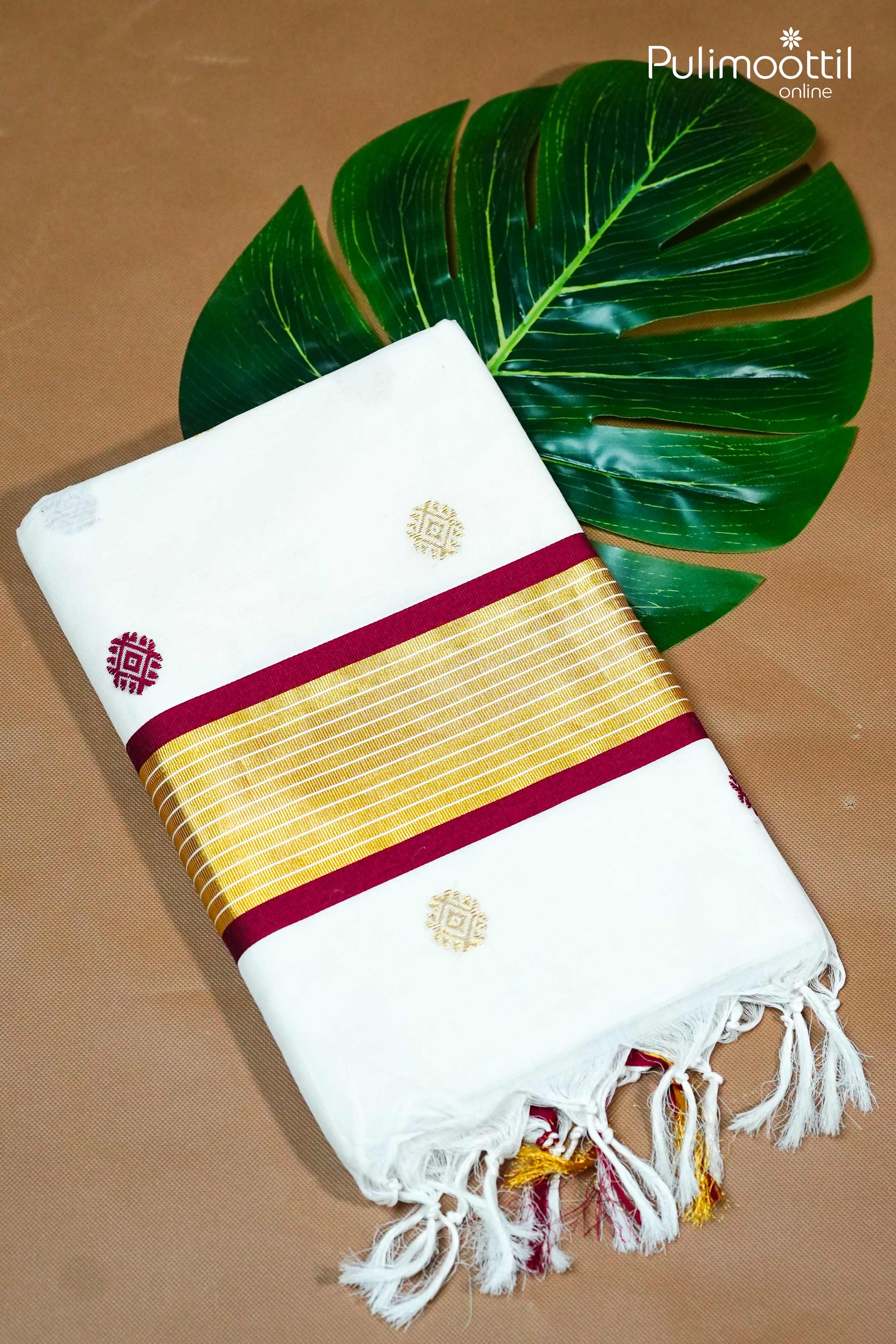 Folded Off White Kerala Set Saree with mixed zari motifs, golden tissue border with maroon edging, and tasselled pallu, placed beside a green tropical leaf.