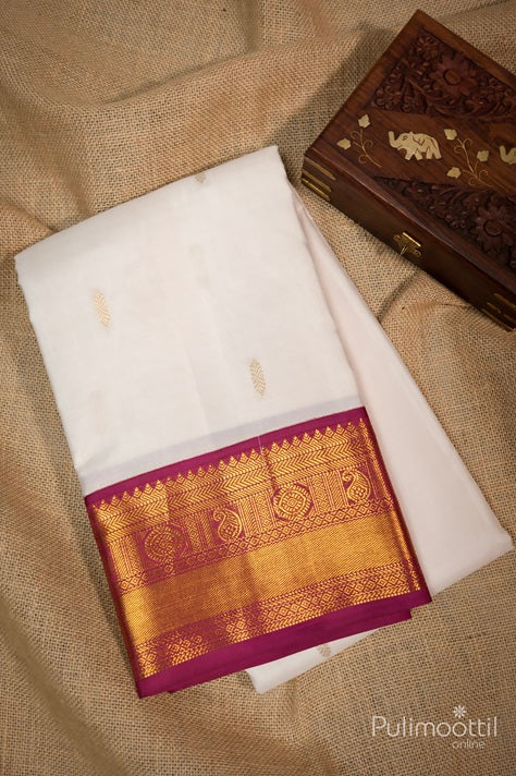 "Off White Pure Kanchipuram Silk Saree with a Golden Zari Border and Maroon Contrast Border, Folded with Decorative Wooden Box in the Background.