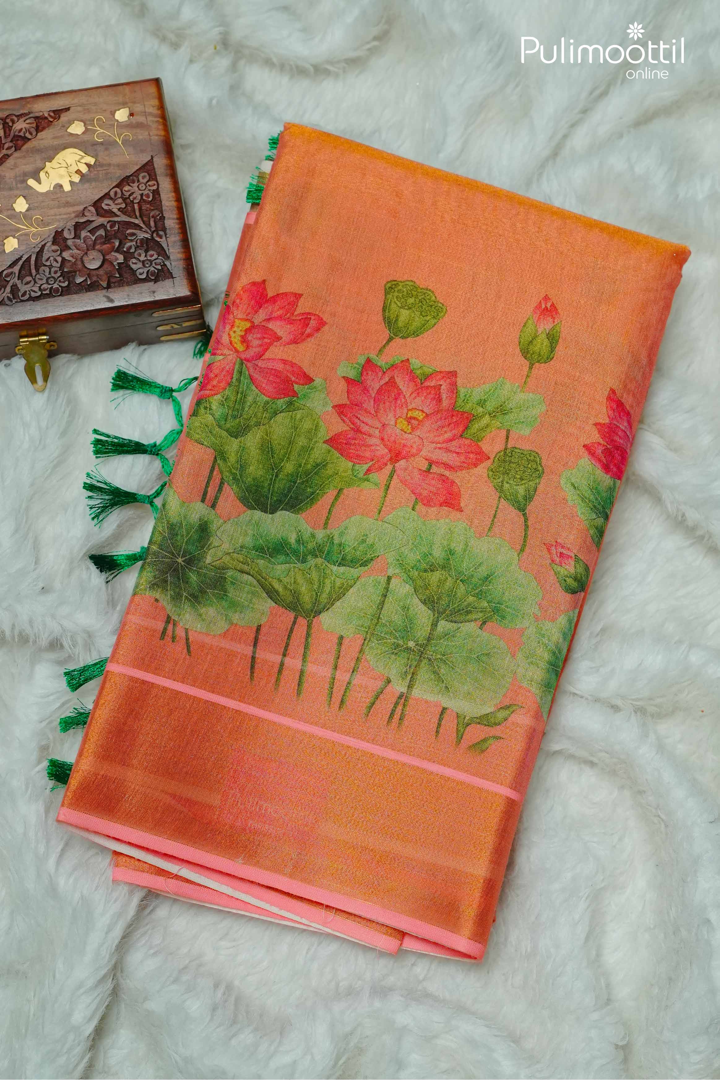 Folded orange Kerala tissue saree with vibrant lotus flower and leaf print on the pallu, finished with green tassels.
