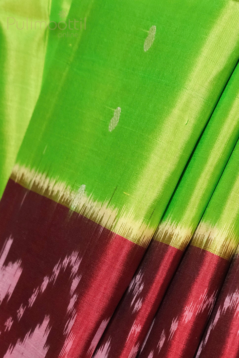 Close-up of a pure soft silk saree showing a vivid parrot-green body with small silver buttas, a gold-tipped transition, and a contrasting maroon ikkat-patterned pallu with white diamond motifs.