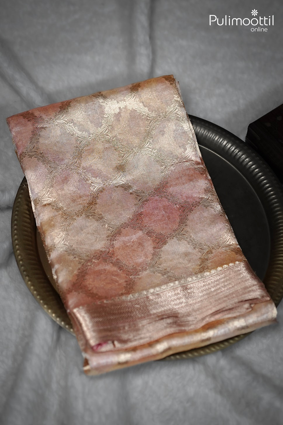 Folded pastel organza silk saree with golden zari border placed on a bronze plate – Pulimoottil Online.