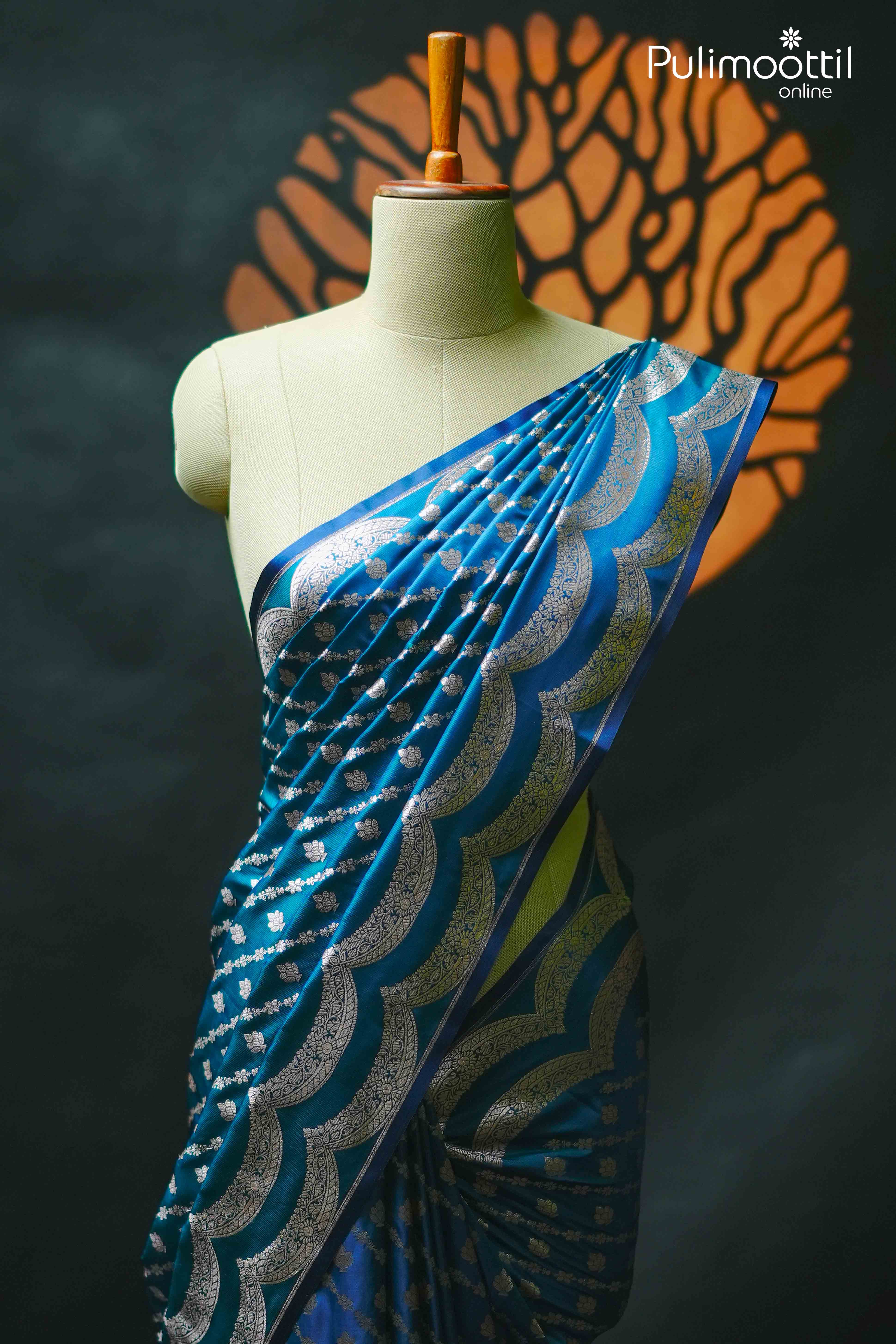 Peacock blue Banarasi silk saree draped on mannequin, featuring silver zari floral buttas and grand scallop border – Pulimoottil Silks Onam Collection
