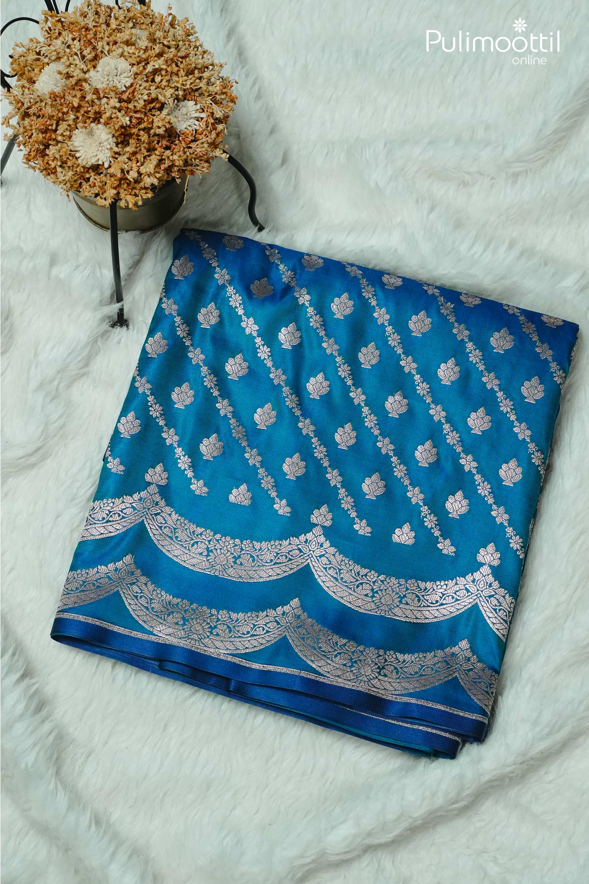 Folded peacock blue Banarasi silk saree with silver zari floral buttas and scalloped border, displayed on soft white background – Pulimoottil Silks
