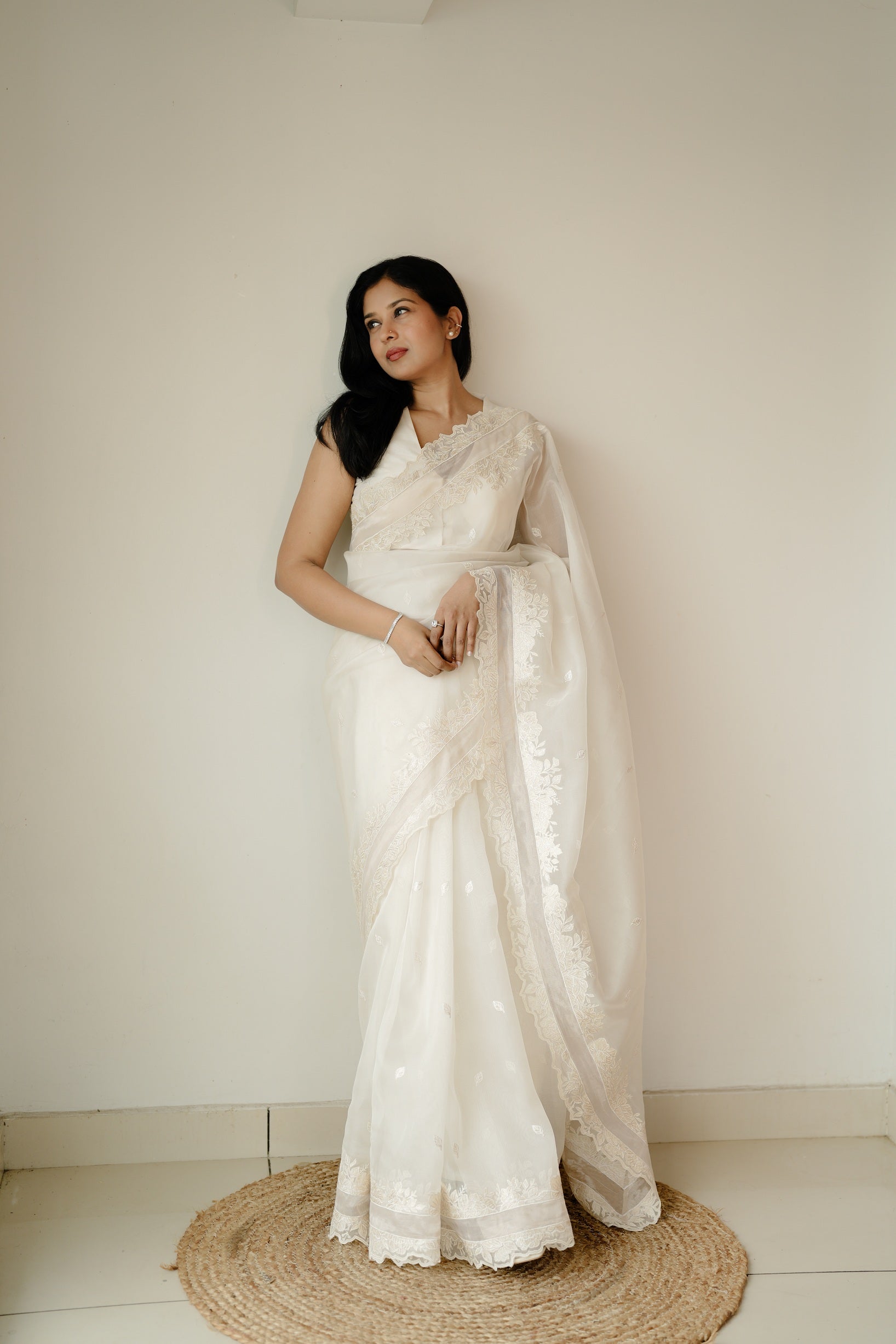 A woman wearing an elegant off-white embroidered saree, standing gracefully on a circular jute rug against a plain cream wall.