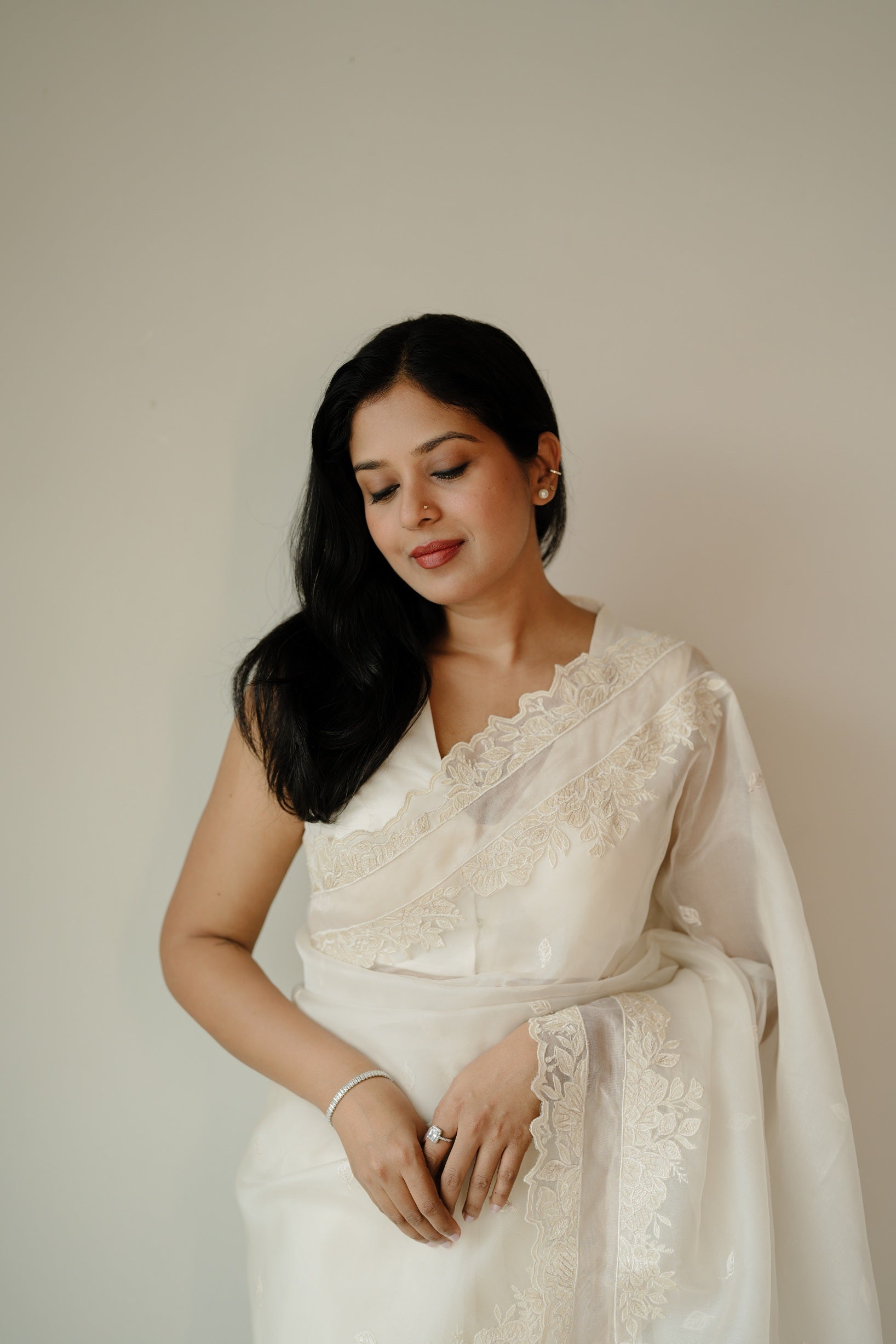 A woman in an off-white saree with delicate floral embroidery, smiling gently with eyes closed, posing against a neutral background.