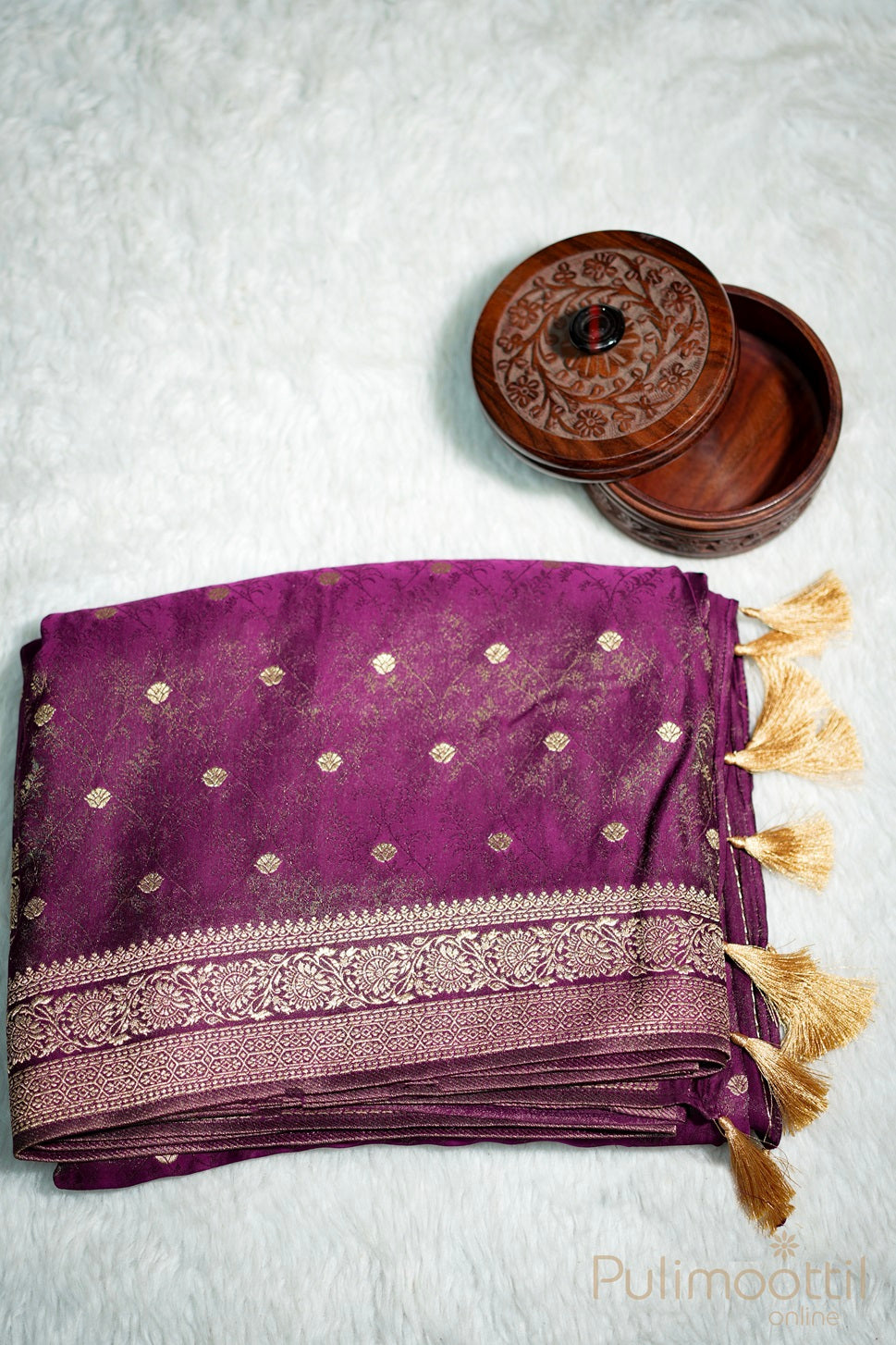 Folded purple Uppada Banarasi silk saree with golden floral buttas, copper zari weave, and tasselled edge, placed beside a carved wooden box.