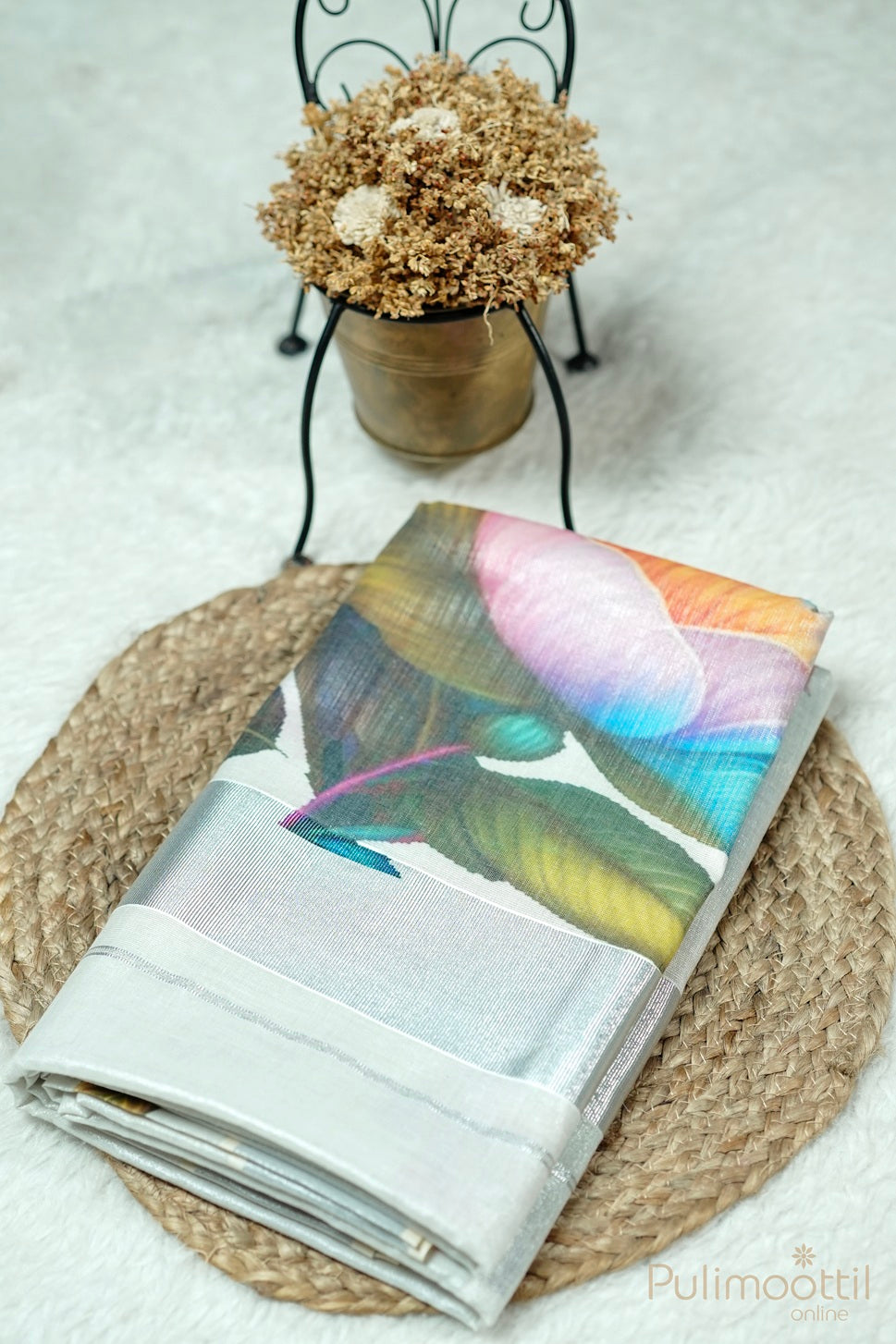 Folded silver tissue Kerala set saree with multicolour floral design and silver border, placed on carpet mat over white fur cloth