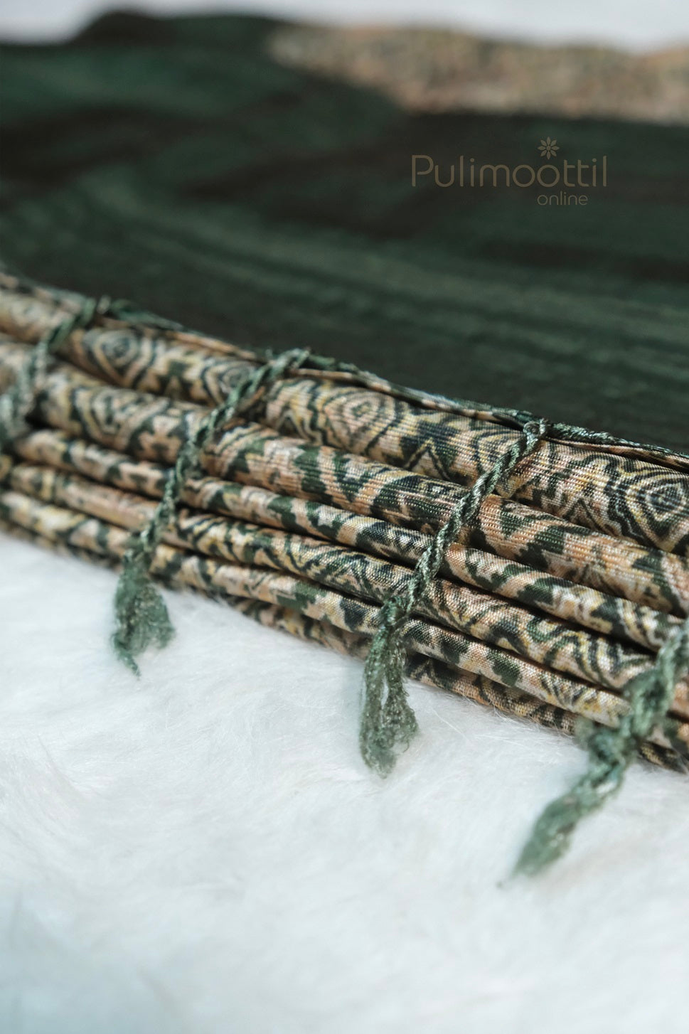 Close-up view of folded green soft silk saree with intricate woven pattern and green tassels on pallu, displayed on white surface with Pulimoottil Online branding.