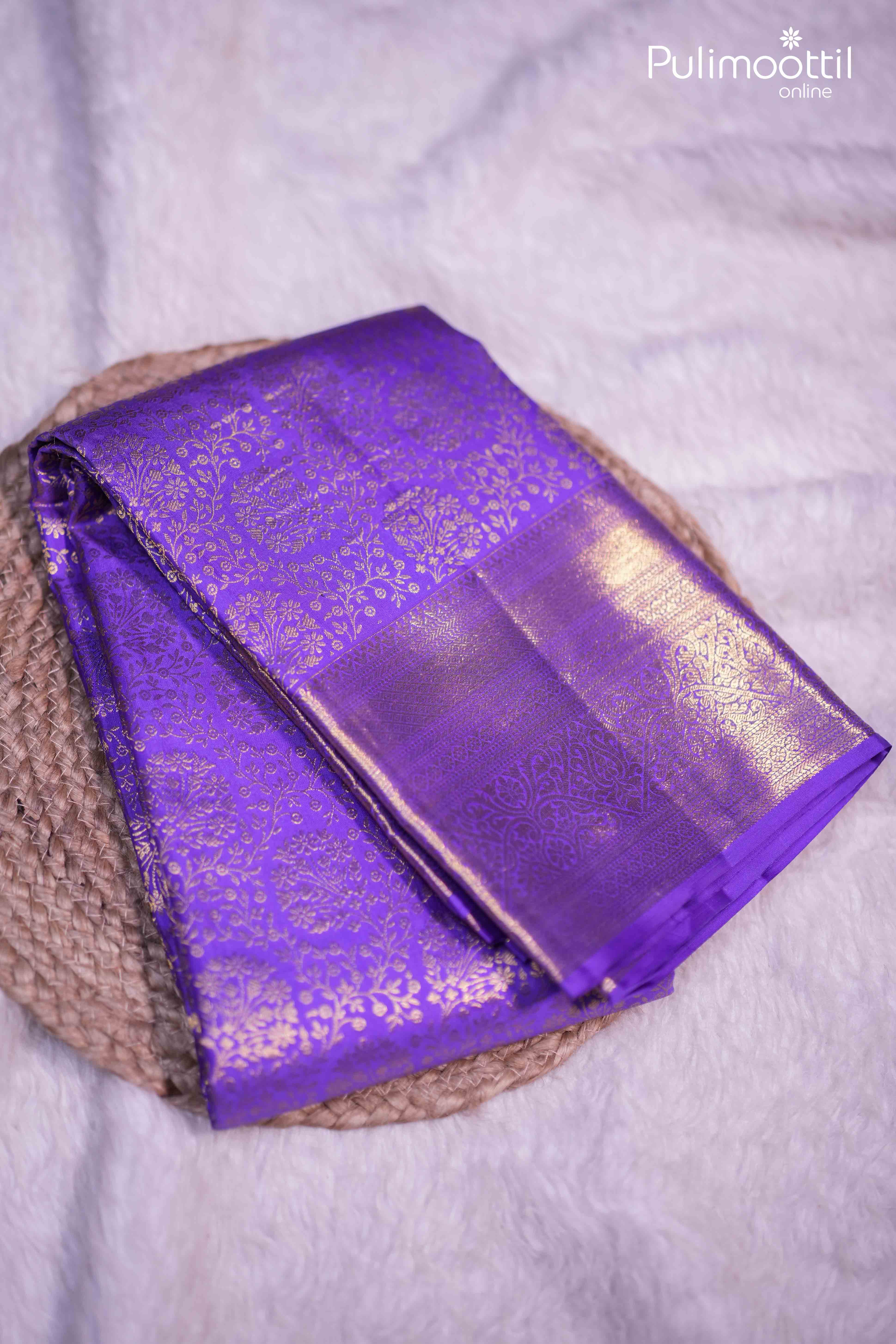Violet Kanchipuram saree with golden zari floral brocade and geometric patterns, neatly folded on a woven mat, showcasing its glossy finish and luxurious design.