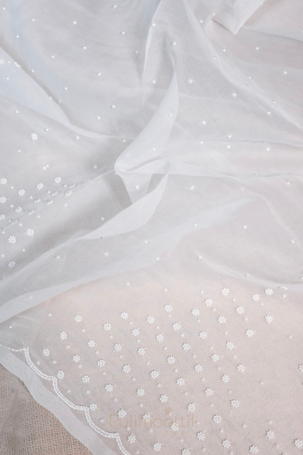 White net saree with scattered pearl and floral embroidery, featuring scalloped beaded border displayed on fabric background