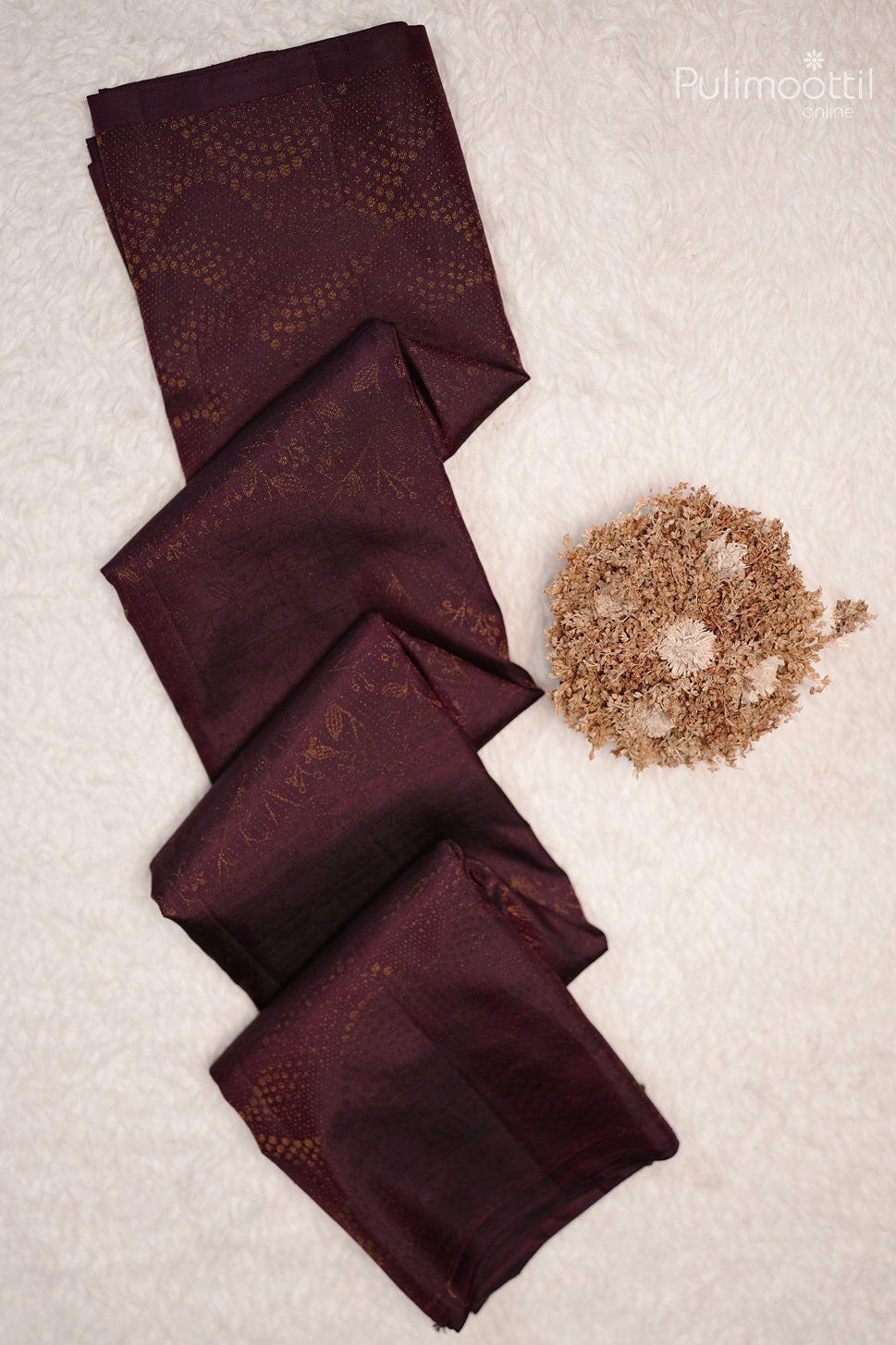 Dark purple Kanchipuram designer saree neatly folded and displayed on a white textured background with a bunch of dried flowers beside it.