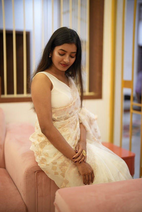 Woman sitting gracefully on a pink velvet sofa wearing a white organza saree with floral pitta work and scallop border.