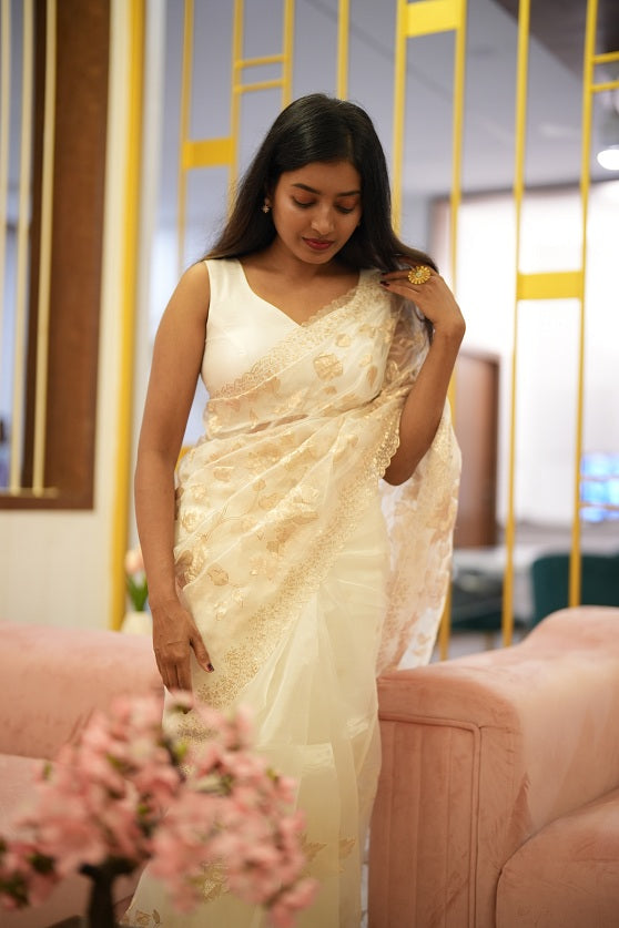 Elegant woman in a white floral organza saree standing in a modern interior setting with golden decor accents.

